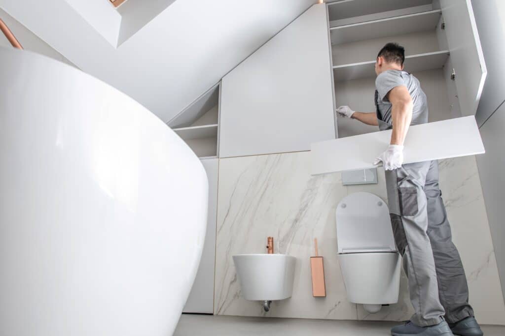 Ouvrier installe meubles blancs dans salle de bain moderne avec baignoire, toilette, bidet et carrelage marbre.