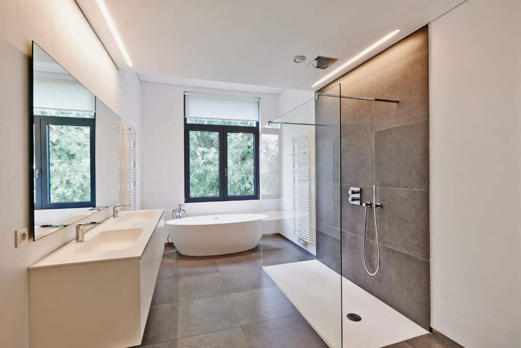 Salle de bain moderne avec baignoire îlot, double vasque miroir, douche à l'italienne carrelée et fenêtre sur verdure. Éclairage LED.